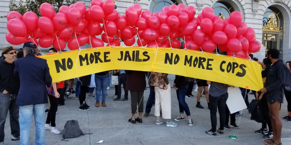 Photo of protestors holding red balloon and sign that reads No More Jails No More Cops